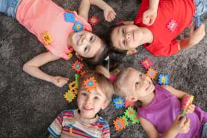 Smiling children lying on a gray carpet, playing with colorful foam number puzzle pieces scattered around them.